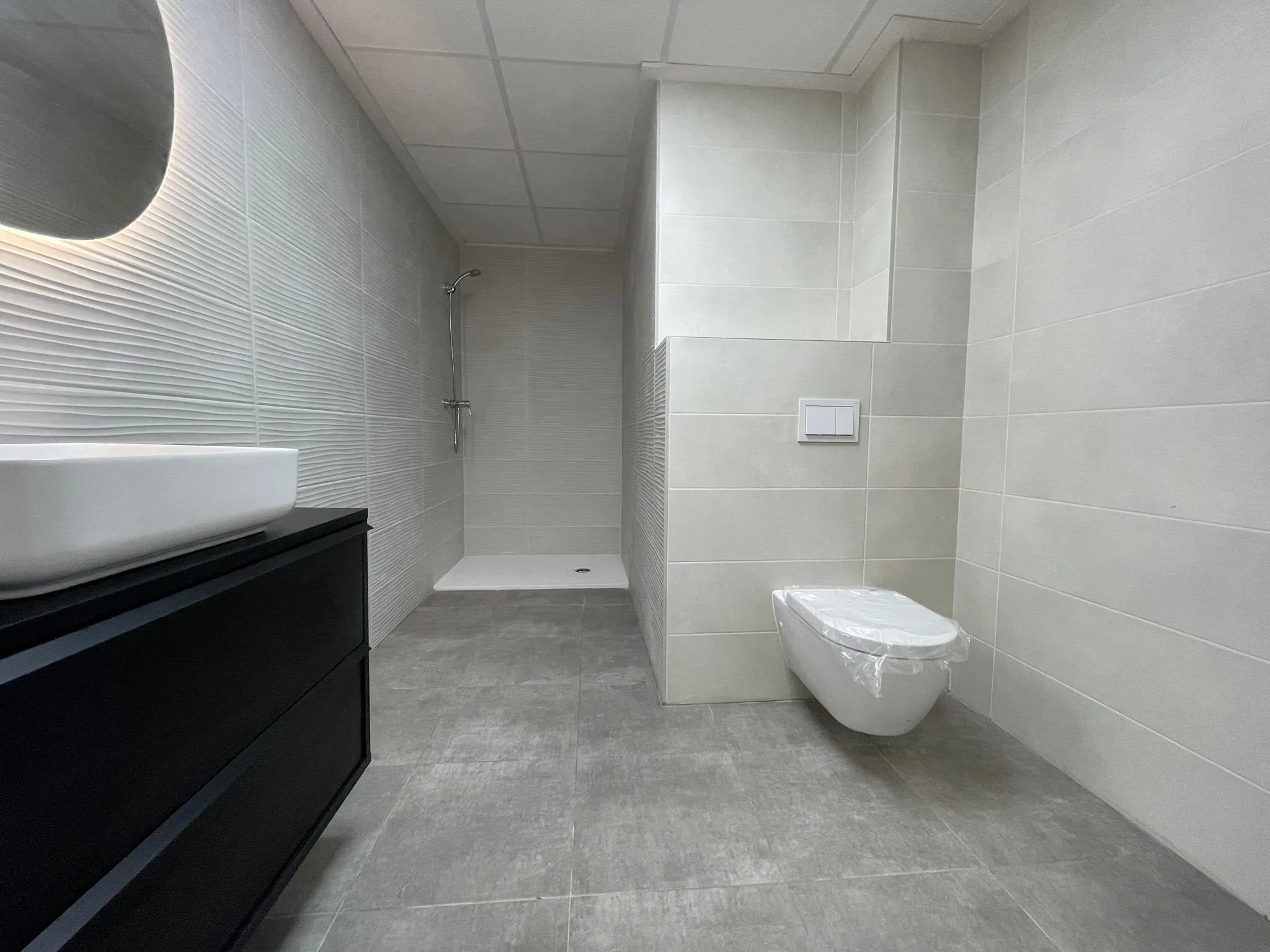 Baño con mueble de lavabo negro, inodoro suspendido y ducha de obra